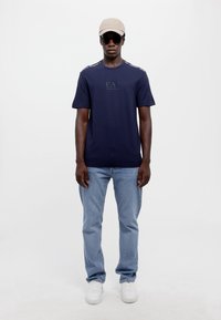 Marineblauw crewneck t-shirt met korte mouwen en subtiele EA7-logo, gecombineerd met lichtblauwe jeans en witte sneakers, accessoires gemaakt met een beige pet en een zonnebril.