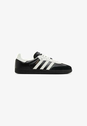 Baskets Adidas Samba en noir et blanc avec trois bandes blanches, semelle noire, lacets blancs et inscription dorée "SAMBA" sur le côté.