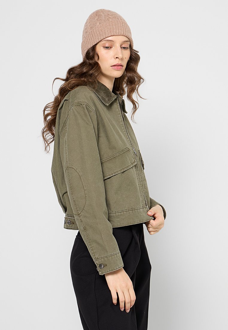 Kobieta z długimi kręconymi włosami, nosząca beżową dzianinową czapkę, oliwkową krótką kurtkę z dużymi przednimi kieszeniami i czarne spodnie, patrzy w dół.