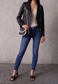 Kvinna iklädd svart läderjacka, krämfärgad ribbad linne, blå slim fit jeans, svarta klacksandaler och bärande på en svart handväska med vit kant.