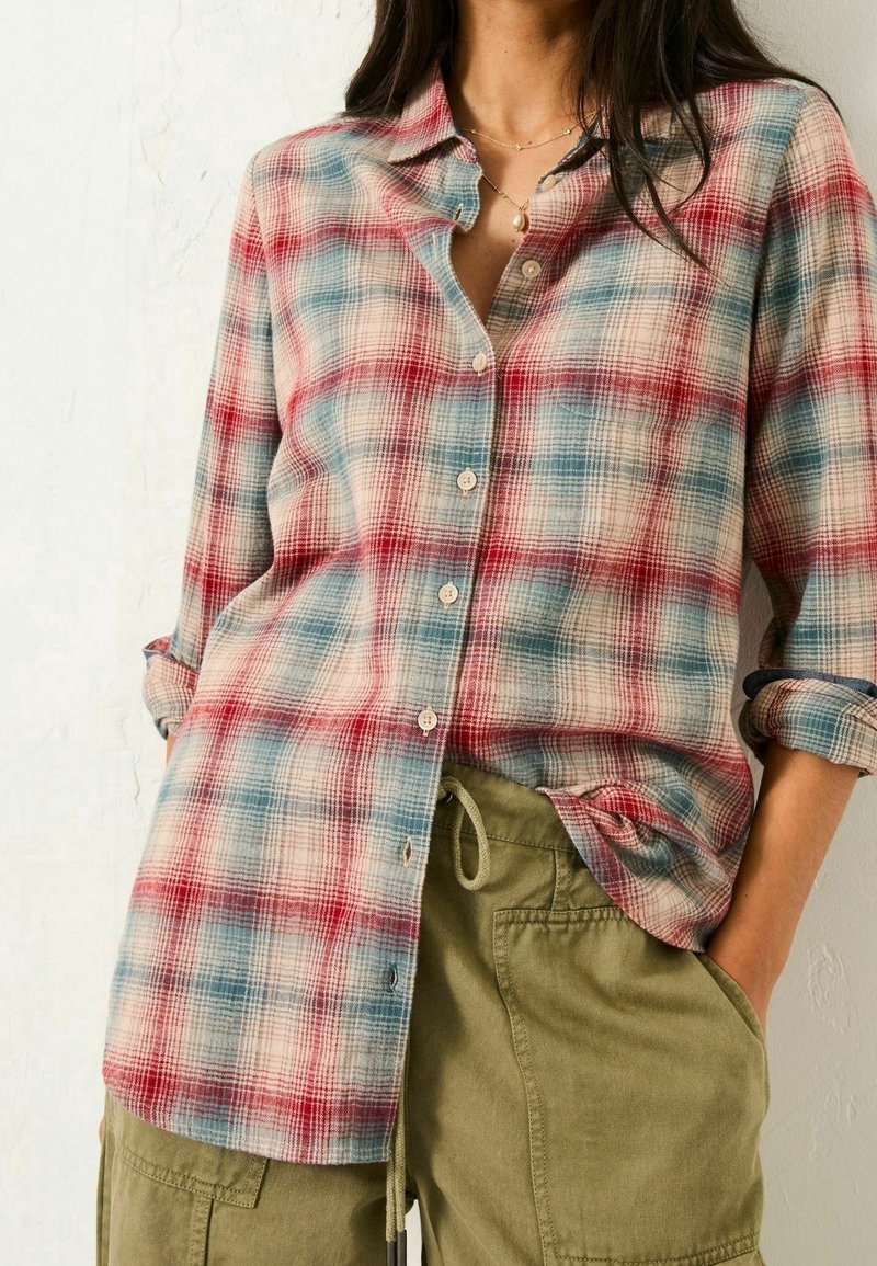 Femme portant une chemise à carreaux rouge et bleue boutonnée avec manches retroussées et un pantalon vert olive à cordon avec poches.
