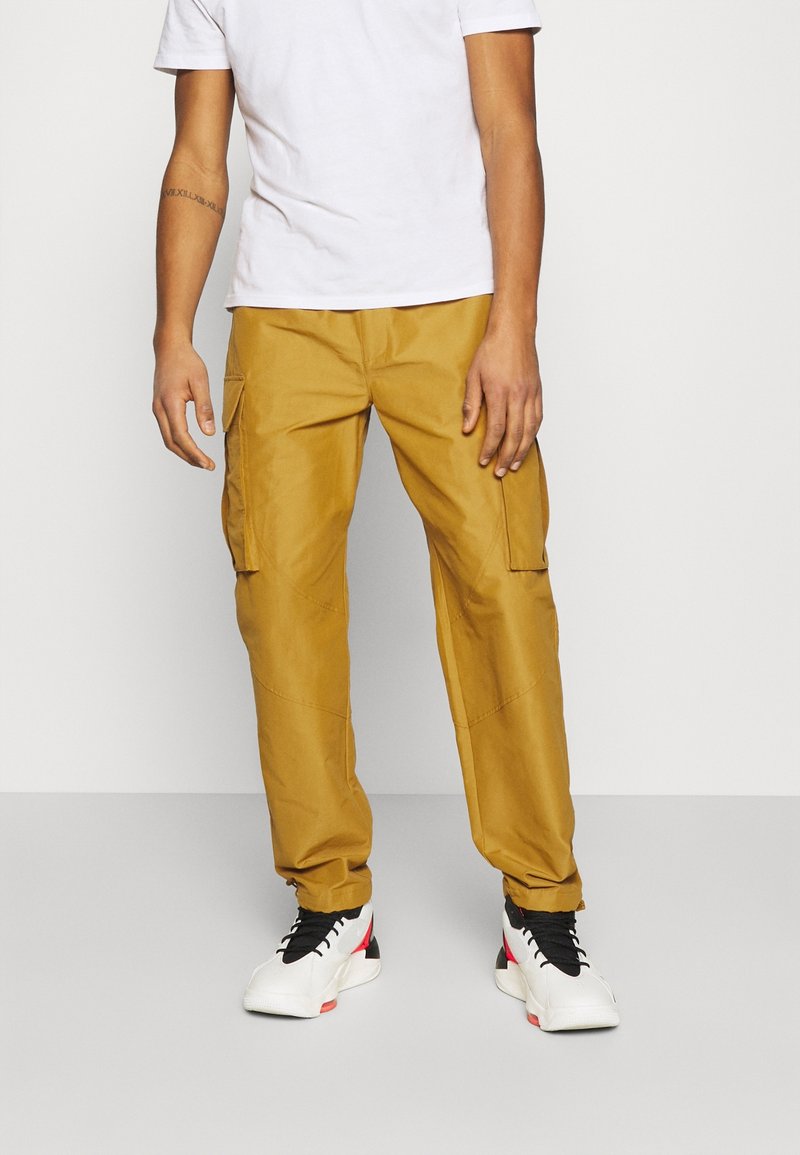 Nákladní kalhoty v zlatavě žluté barvě, vyrobené z pevné látky. Mají boční kapsy a volný střih. Kombinovány s bílými a červenými teniskami.