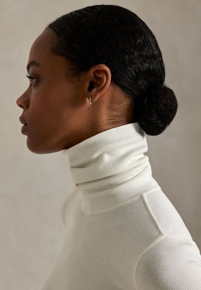 Profilkép egy sötét hajú nőről, alacsony kontyban, fehér bordázott garbóval és kis arany karika fülbevalóval, semleges háttér előtt.