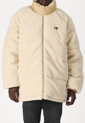 Homme portant une veste polaire couleur crème avec un col beige et un petit logo rectangulaire sur la poitrine, associé à un pantalon foncé.