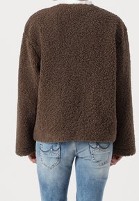 En person som bär en brun, strukturerad fleecejacka och ljusblå denimjeans, sedd bakifrån mot en enhetlig bakgrund.