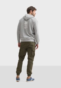 Un sweat à capuche gris avec un imprimé au dos, un pantalon cargo vert avec des poches latérales, et des chaussures de sport bleues avec des accents marron, vu de l'arrière.
