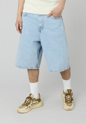 Shorts en denim bleu clair avec une coupe décontractée, présentant des coutures surpiquées et une texture lisse. Associés à des baskets beiges et brunes et des chaussettes blanches.