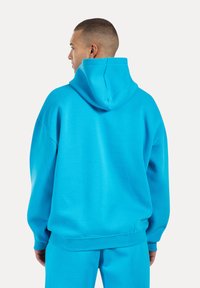 Sudadera con capucha de color turquesa en tejido suave, que presenta un corte relajado y puños de canalé, con un bolsillo tipo canguro y detalles de costura.