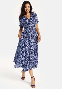 Blue and white patterned maxi dress with short sleeves, smocked waist, and side pockets, paired with blue heeled sandals.