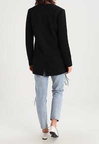 Chaqueta negra de manga larga con tejido texturizado, espalda dividida, combinada con jeans azul claro y zapatillas blancas con acento negro.