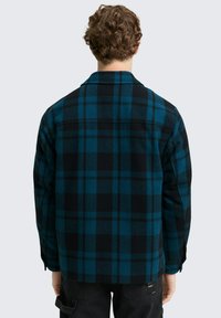 Blauw en zwart geruit overhemd met een kraag, lange mouwen en een gestructureerde wollen stof. Heeft een rechte snit en knoopmanchet.