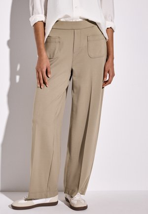 Person trägt beige weit geschnittene Hose mit aufgesetzten Taschen vorne, weiße langärmelige Bluse mit aufgekrempelten Ärmeln und weiße Turnschuhe mit beigen Akzenten.