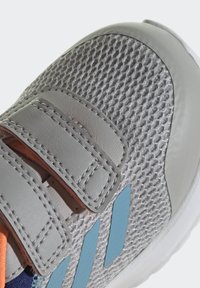 Chaussure de sport grise avec une tige en maille, accents synthétiques argentés, rayures bleues et bandes Velcro orange. Présente une surface texturée et un bout arrondi.