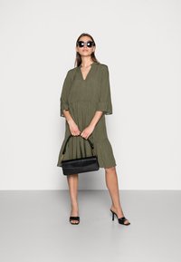 Vestido verde-azeitona, até aos joelhos, com decote em V e mangas três quartos, apresentando um design em camadas. A modelo segura uma mala preta com detalhes em dourado.