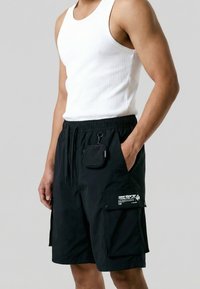 Άνδρας που φοράει μαύρα cargo σορτς με μια μικρή μαύρη τσάντα και ένα λευκό αμάνικο ριμπ τοπ.