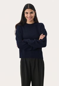 Pull en maille bleu marine avec un col rond côtelé et des coutures d'épaule tombantes, associé à un pantalon noir à taille haute.