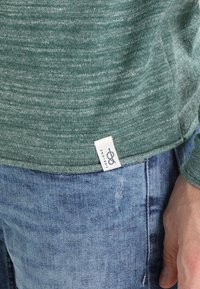 Grön stickad tröja med vit Pier One-etikett instoppad i ljusblå denimjeans, hand vilande nära fickan.