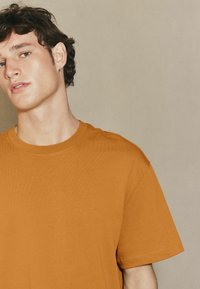 Kurzarm-T-Shirt mit Rundhalsausschnitt in einer strukturierten Bernsteinfarbe, mit einer lässigen Passform und sichtbaren Nahtdetails an den Ärmeln und dem Ausschnitt.