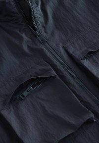 Jachetă neagră ușoară cu fermoar frontal, două buzunare cu fermoar și o textură crăpată. Dispune de un guler înalt și un design minimalist.