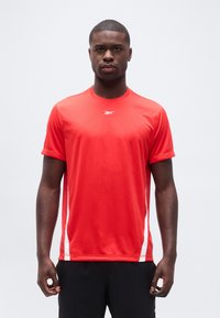 Άνδρας που στέκεται με το πρόσωπο προς τα εμπρός, φορώντας κόκκινο αθλητικό κοντομάνικο μπλουζάκι με λευκές πλαϊνές ρίγες και μαύρο σορτς, στο φόντο ενός απλού σκηνικού.