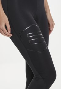 Svarta leggings tillverkade av elastiskt tyg, med ett glänsande svart geometriskt mönster på låret och en åtsittande silhuett.