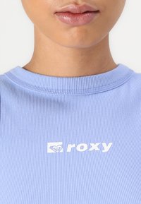 Lys lilla ribbet t-skjorte med rund hals, med hvit logo tekst "roxy" i midten, som fremhever jevn tekstur og subtile detaljer.