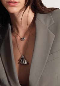 Collares en tono plateado con colgantes únicos y escultóricos; uno pequeño, en forma de lágrima, y el otro más grande, que se asemeja a tela que fluye.