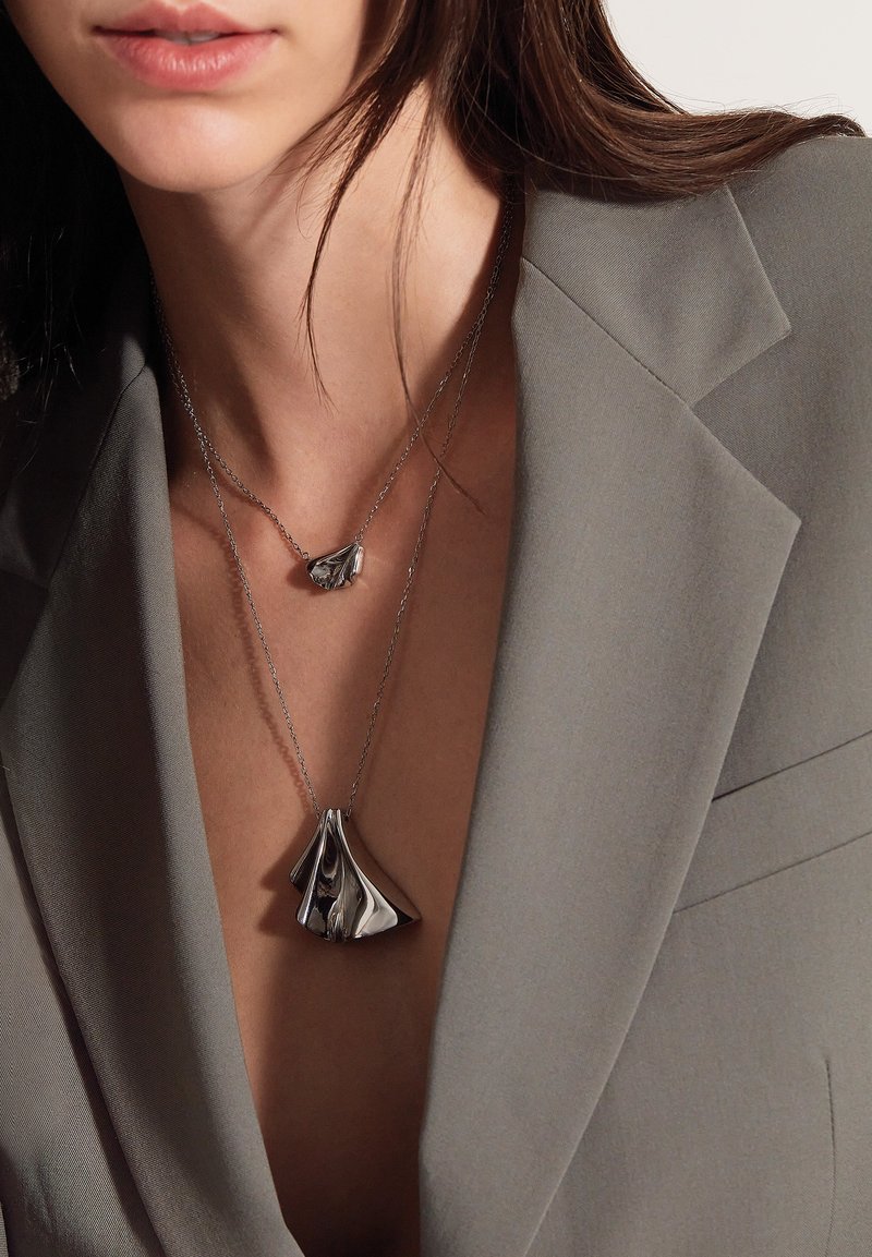 Collares en tono plateado con colgantes únicos y escultóricos; uno pequeño, en forma de lágrima, y el otro más grande, que se asemeja a tela que fluye.
