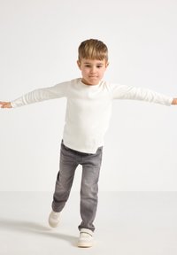 Hvid langærmet skjorte med ribbet tekstur, parret med grå jeans. Barnet står med armene strakt ud, iført lyse sko.