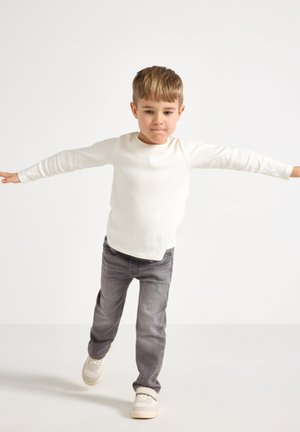 Camicia bianca a maniche lunghe con una texture a coste, abbinata a jeans grigi. Il bambino è in piedi con le braccia tese, indossando scarpe chiare.