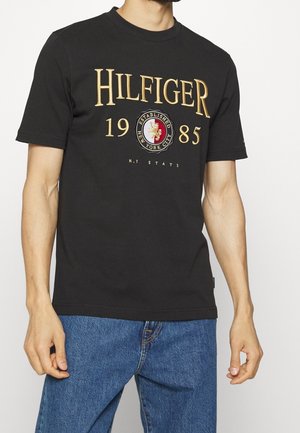 Mężczyzna w czarnej koszulce Hilfiger ze złotym napisem ""HILFIGER 1985"", logo z herbem i niebieskimi dżinsami, pokazany od ramienia do uda.