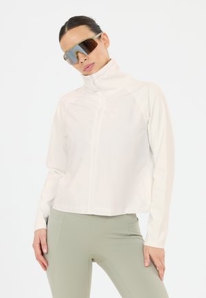 Woman wearing white zip-up jacket, light green leggings, and reflective oversized sunglasses, standing with hands near hips.