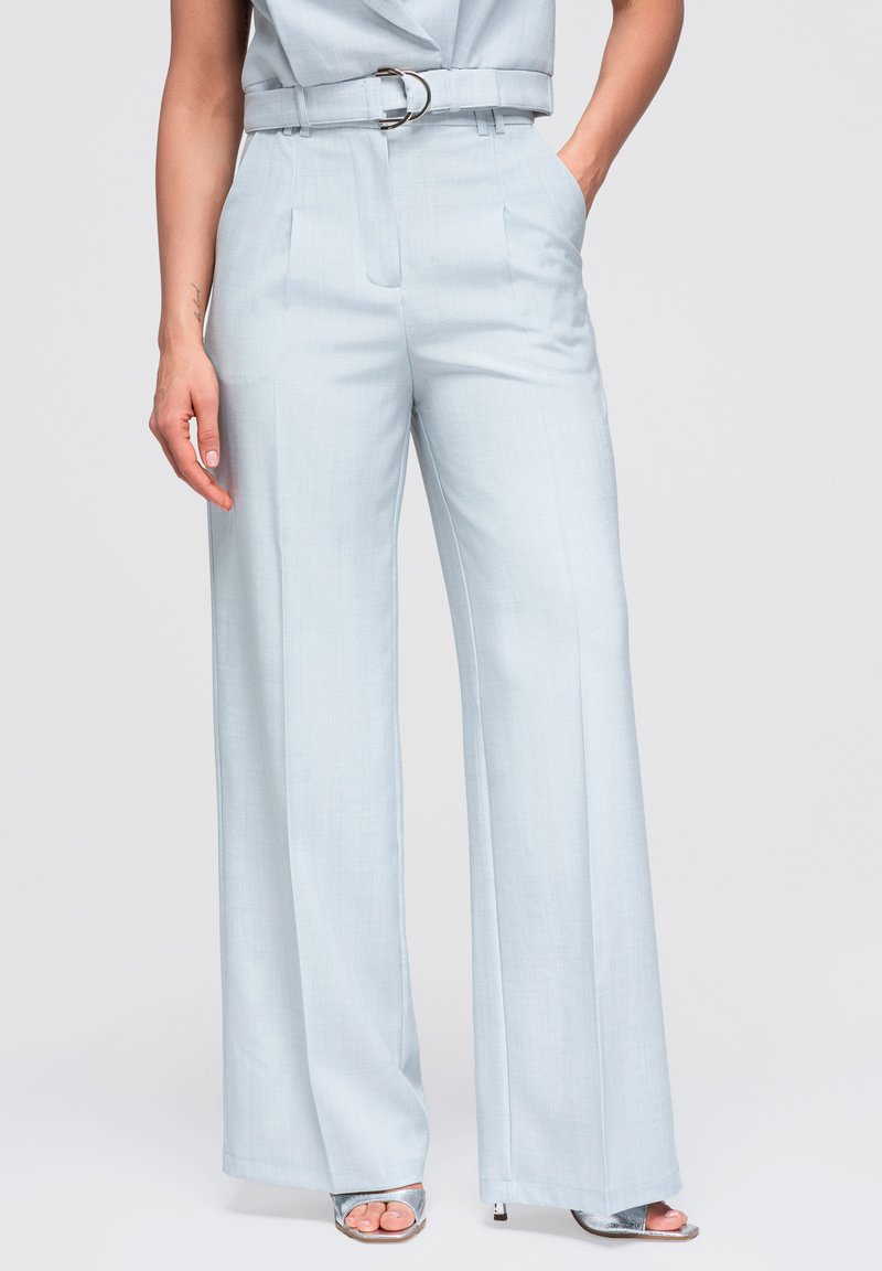Femme portant un pantalon taille haute bleu clair à jambes larges avec ceinture et des sandales à talons hauts argentées, debout avec une main dans la poche.