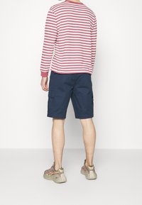 Osoba stojící čelem ke zdi, oblečená v tričku s dlouhým rukávem červeno-bílých pruhů, námořnických šortkách a béžovo-růžových teniskách.