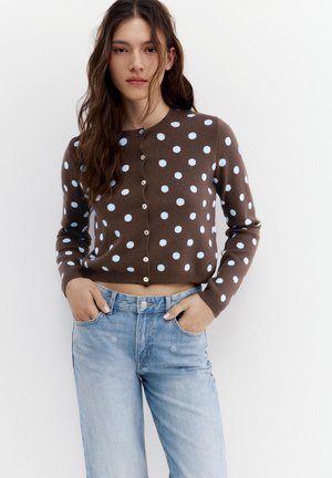 Młoda kobieta z długimi falującymi włosami, ubrana w brązowy krótki kardigan z jasnoniebieskimi kropkami oraz jasnoniebieskie, wysokowalne dżinsy.