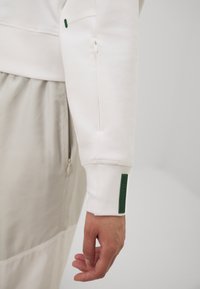 Hanorac alb cu un buzunar cu fermoar pe mânecă, manșete cu textură ribbed și un accent verde texturat. Material moale cu un finisaj neted.