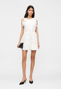 Γυναίκα που στέκεται φορώντας λευκό φόρεμα με βολάν και ανοιχτούς ώμους, μαύρες γόβες και κρατώντας μαύρο τσαντάκι, μπροστά σε μονόχρωμο φόντο.