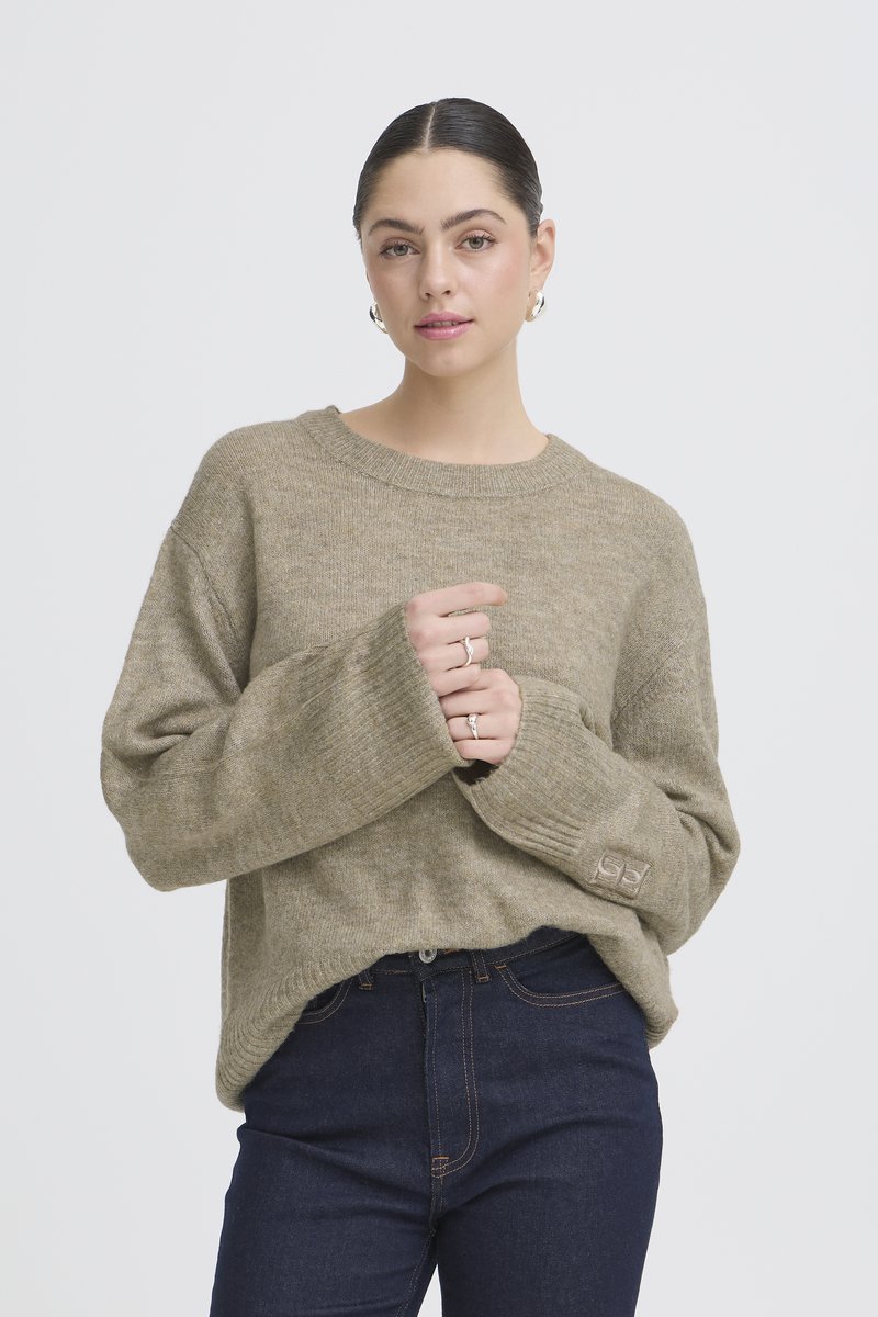 Jeune femme aux cheveux foncés portant un pull beige oversize et un jean foncé, debout les mains jointes près de la poitrine devant un fond uni.