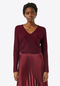 Burgundowy sweter w serek z miękkiego materiału, w połączeniu z plisowaną midi spódnicą w dopasowanym odcieniu, tworzy spójną monochromatyczną stylizację.