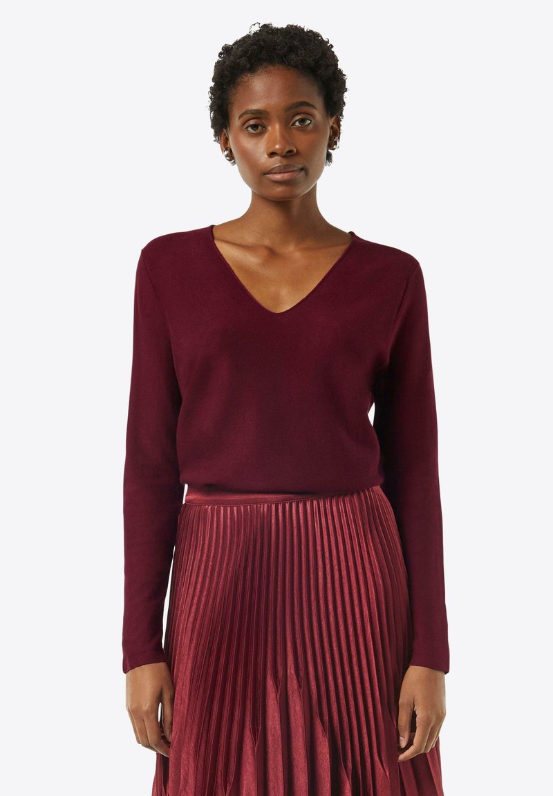 Burgundowy sweter w serek z miękkiego materiału, w połączeniu z plisowaną midi spódnicą w dopasowanym odcieniu, tworzy spójną monochromatyczną stylizację.