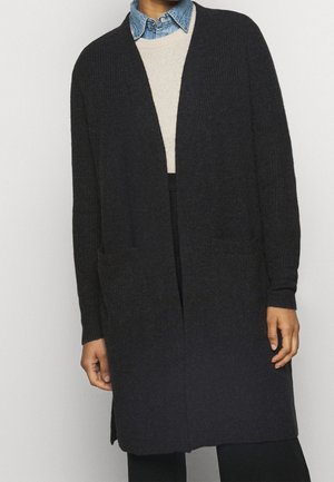 Personne portant un long cardigan gris foncé ouvert sur un pull beige et une chemise en denim bleu à col.