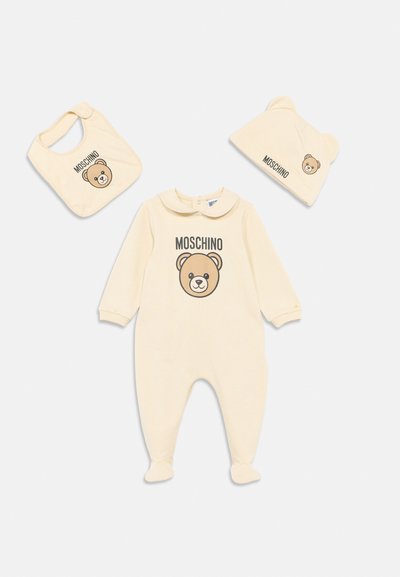 Conjunto para bebé color crema que incluye un body con pies, un babero y un gorro, cada uno con la cara de un osito de peluche y la marca "Moschino".