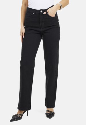 Vrouw draagt zwarte high-waist rechte jeans, witte top, zilveren armband en zwarte puntige pumps met enkelbandjes.