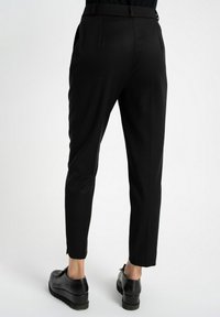 Czarne zwężane spodnie o gładkiej teksturze, z dopasowanym pasem, bocznymi kieszeniami i subtelnymi fałdami. Noszone z czarnymi butami.