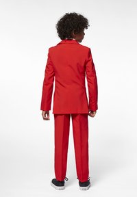Rode pak met bijpassende broek, met een nauwkeurige pasvorm. De jas heeft een gesneden kraag en een gladde textuur. Witte binnenvoering zichtbaar bij de kraag.