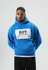 Sweat à capuche bleu avec une grande étiquette blanche affichant le chiffre "009" et du texte. Le tissu semble doux, avec une poche avant et des détails texturés.