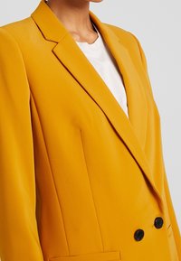 Dwurz�ędowa żółta marynarka wykonana z gładkiej tkaniny, z ostrym kołnierzem, ozdobiona dwoma czarnymi guzikami i bocznymi kieszeniami.