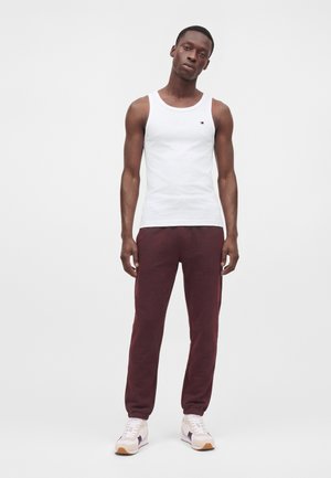 Homme portant un débardeur blanc, un pantalon de jogging bordeaux foncé et des baskets blanches, debout devant un fond blanc uni.