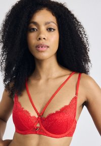 Red lace bra with contrast detailing. Features intricate floral patterns, adjustable, textured straps, and a decorative keyhole at the center.