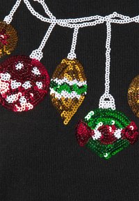 Schwarzer Pullover, geschmückt mit bunten Paillettenornamenten in Rot, Gold und Grün, die verschiedene Formen und Muster aufweisen und auf einem weißen Faden aufgereiht sind.